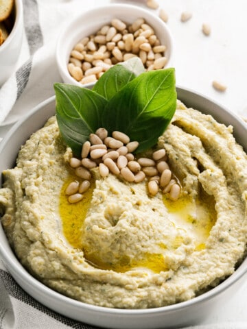 A bowl of pesto hummus garnished with a drizzle of olive oil, pine nuts, and basil leaves.