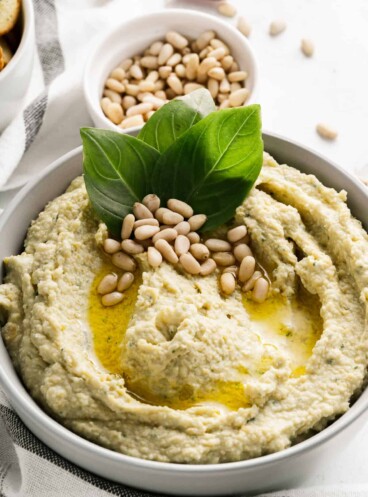 A bowl of pesto hummus garnished with a drizzle of olive oil, pine nuts, and basil leaves.