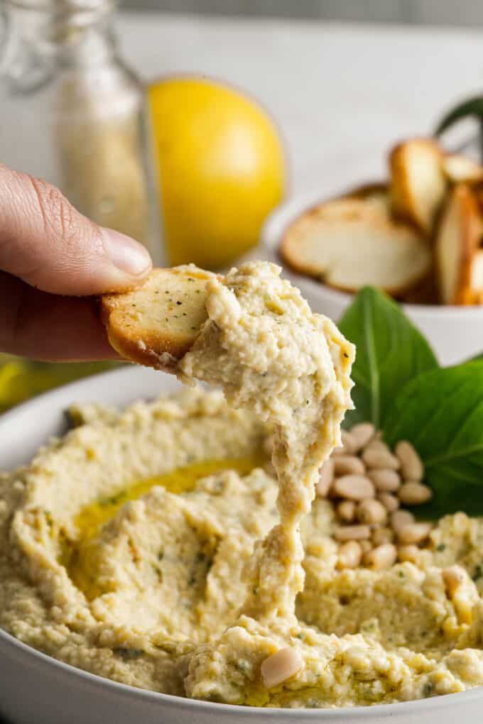 A crunchy bagel chip covered in pesto hummus being lifted out of a bowl.