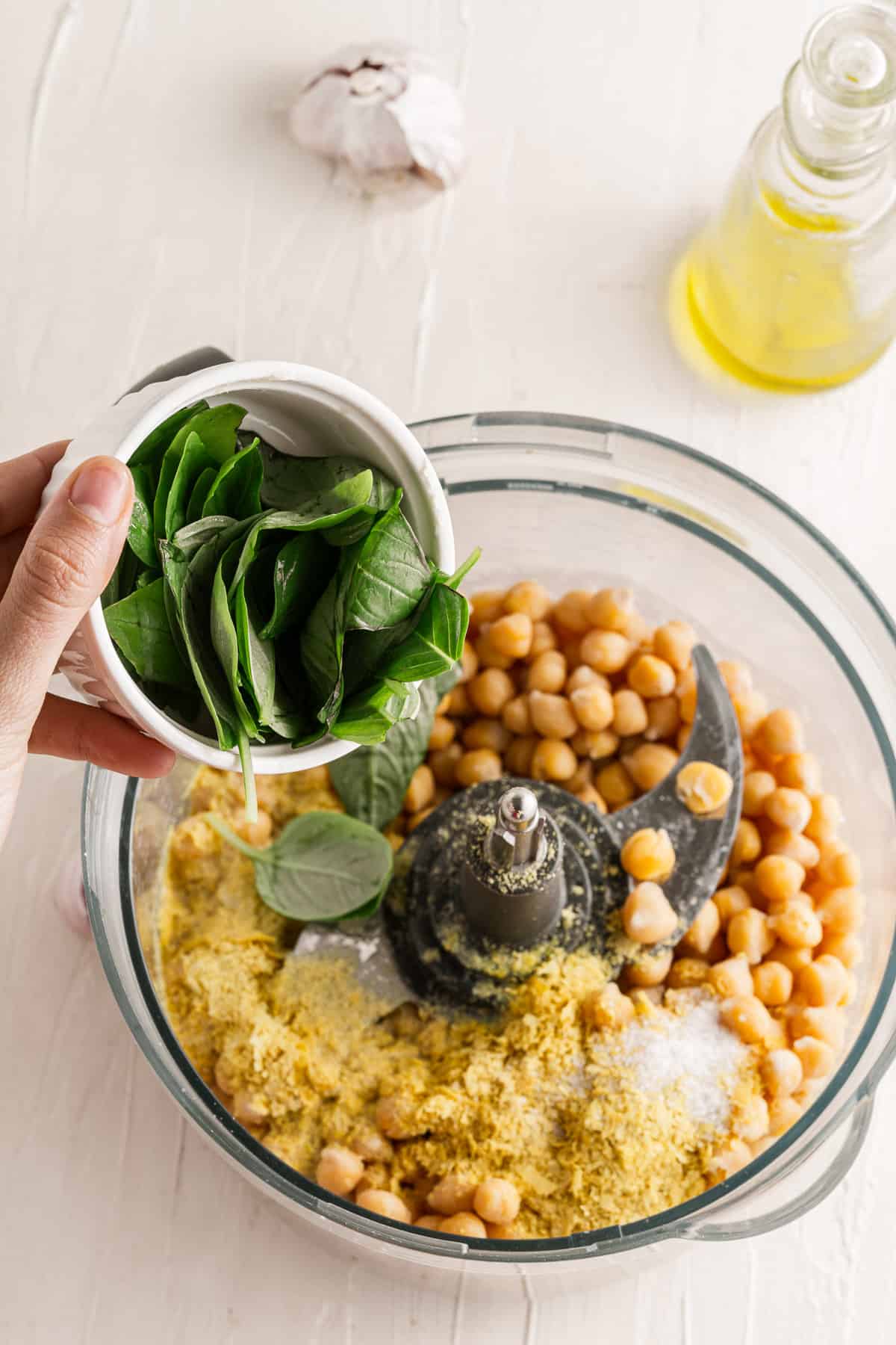 Adding fresh basil leaves to hummus ingredients in a large food processor.