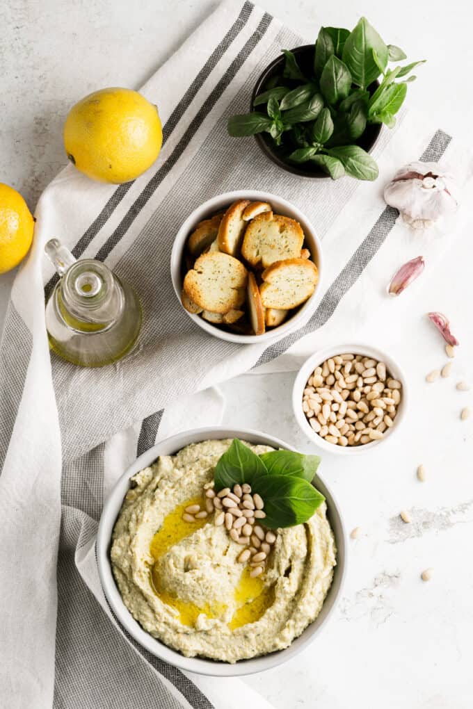 Homemade pesto hummus next to bowls of pine nuts, toasted bagel chips, and fresh basil.