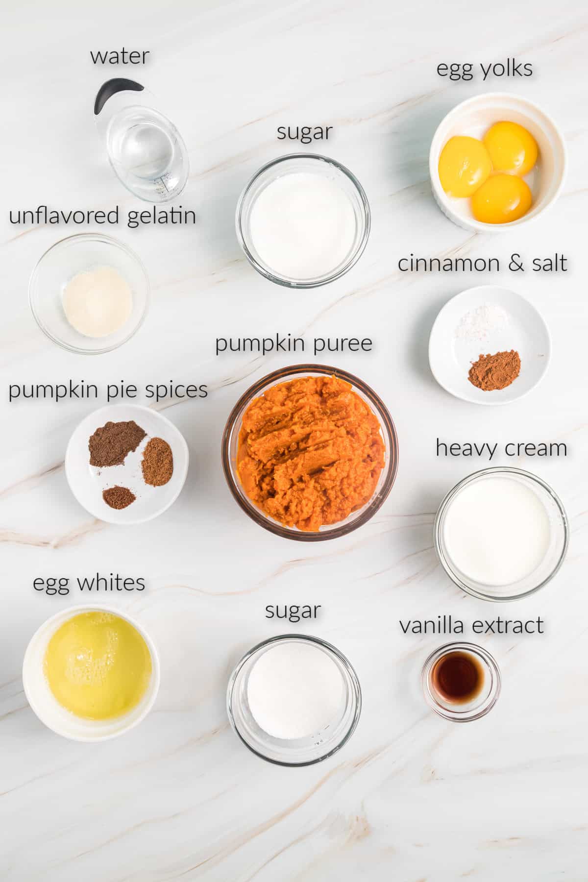 Top view of pumpkin chiffon pie ingredients in bowls including water, sugar, egg yolks, pumpkin puree, cinnamon and salt, gelatin, pumpkin pie spices, heavy cream, egg whites, and vanilla extract. 