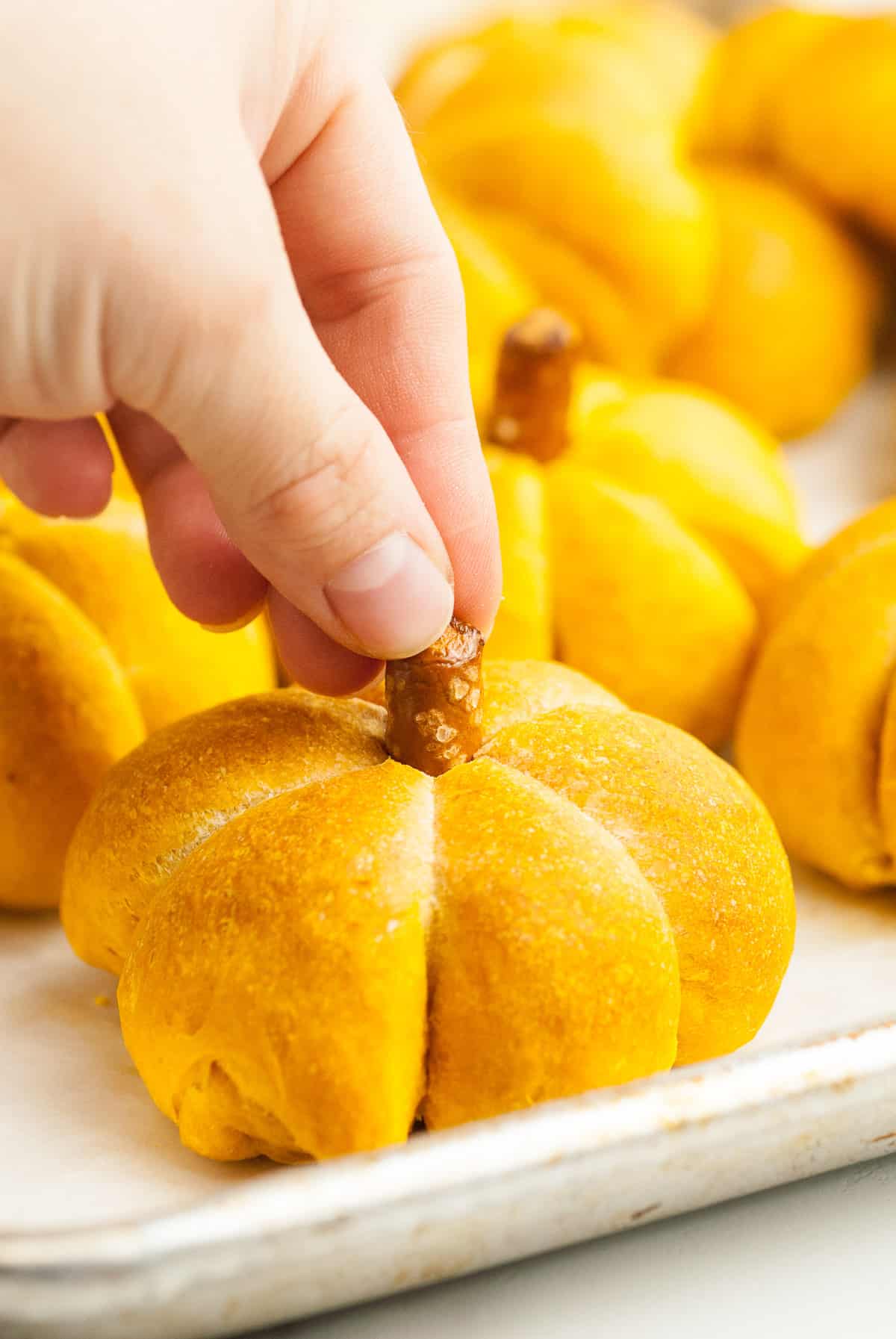 A hand is placing a piece of a pretzel rod in the top center of a pumpkin roll. The twine has been removed. 