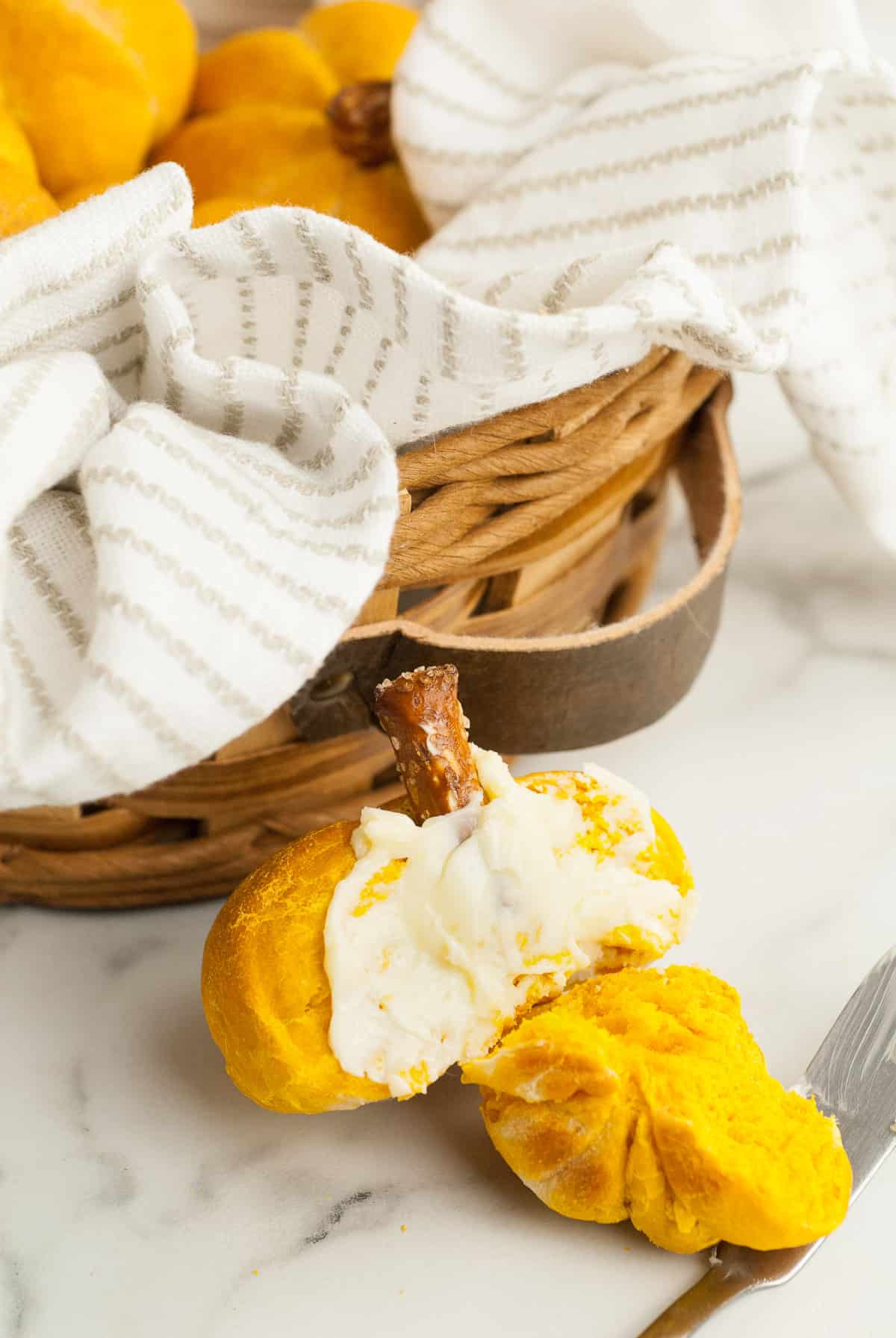 A pumpkin dinner roll is broken apart and slathered with butter, next to a bread basket filled with pumpkin rolls. 