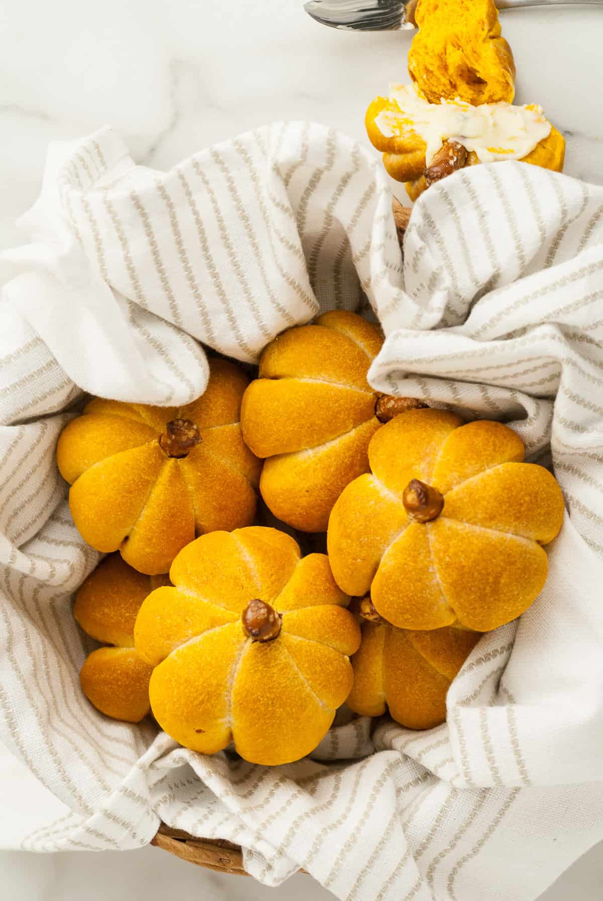 A bread basket lined with a striped dish towel holds multiple pumpkin rolls. 