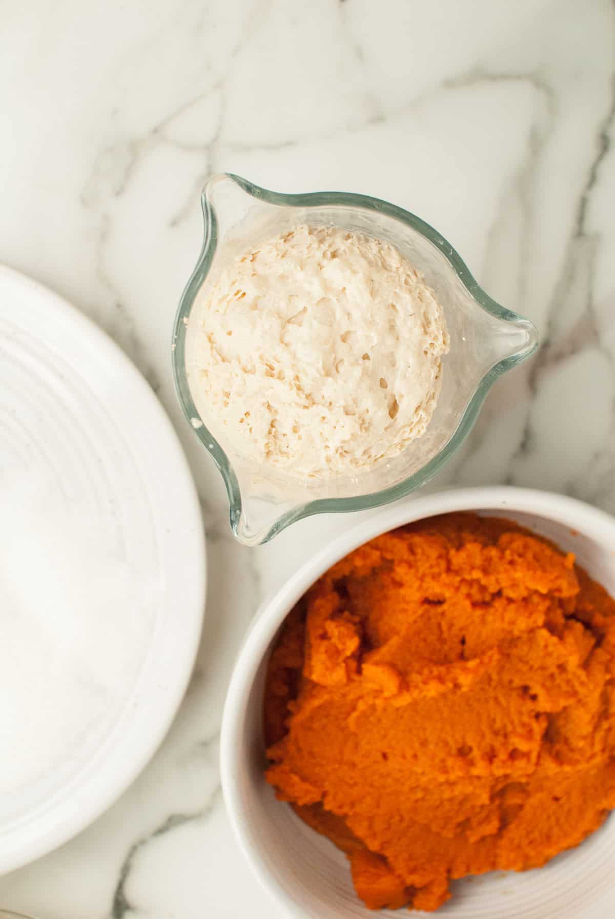 Proofed yeast is sitting in a clear container, next to a bowl of pumpkin puree and other ingredients for pumpkin rolls. 