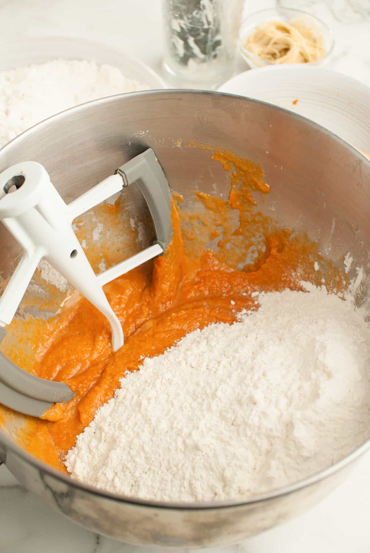 A paddle attachment is resting in a silver metal bowl. Pumpkin puree and dry ingredients are visible in the bowl. 