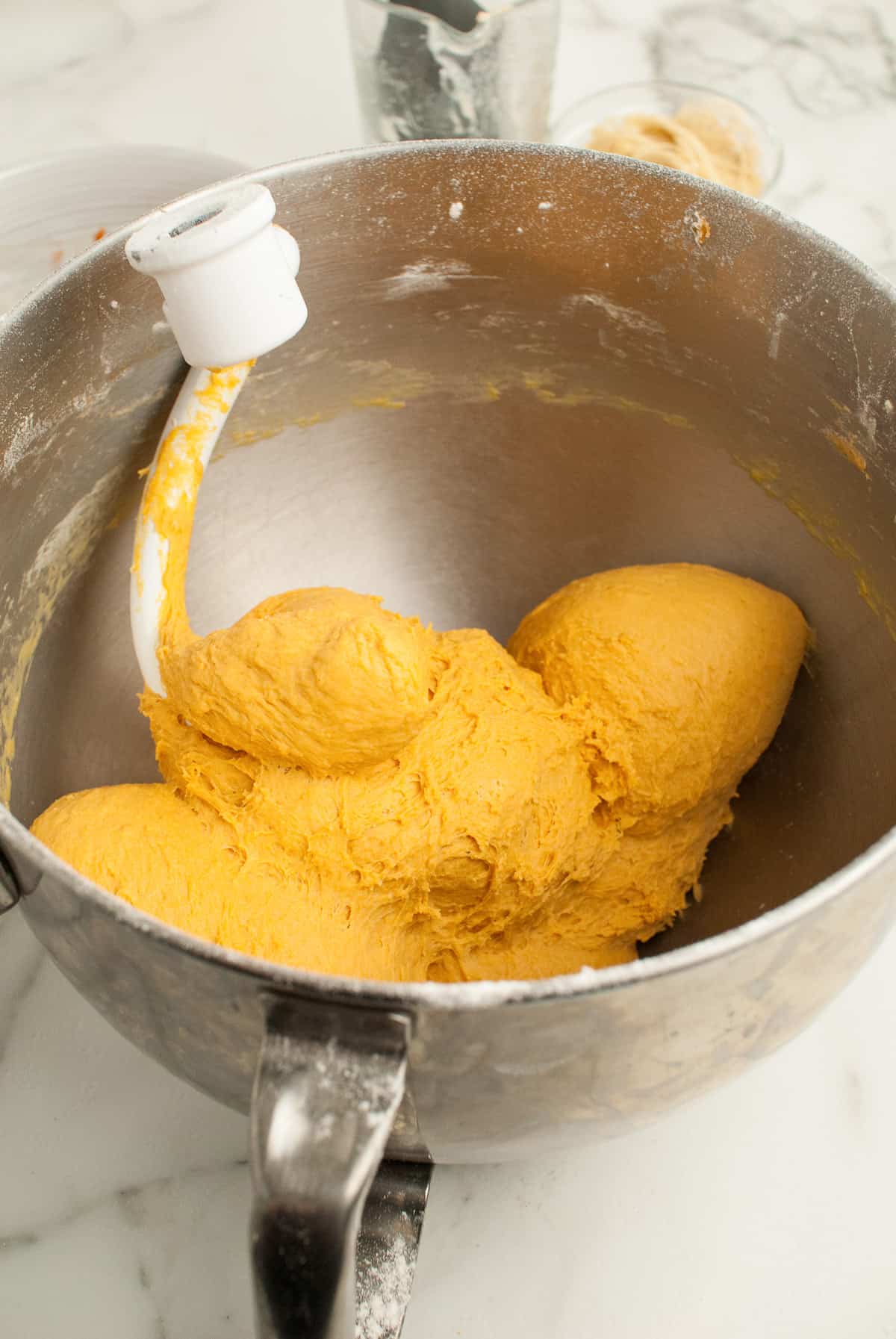 Pumpkin roll dough is resting in a silver metal bowl. A dough hook is sitting with the dough. 