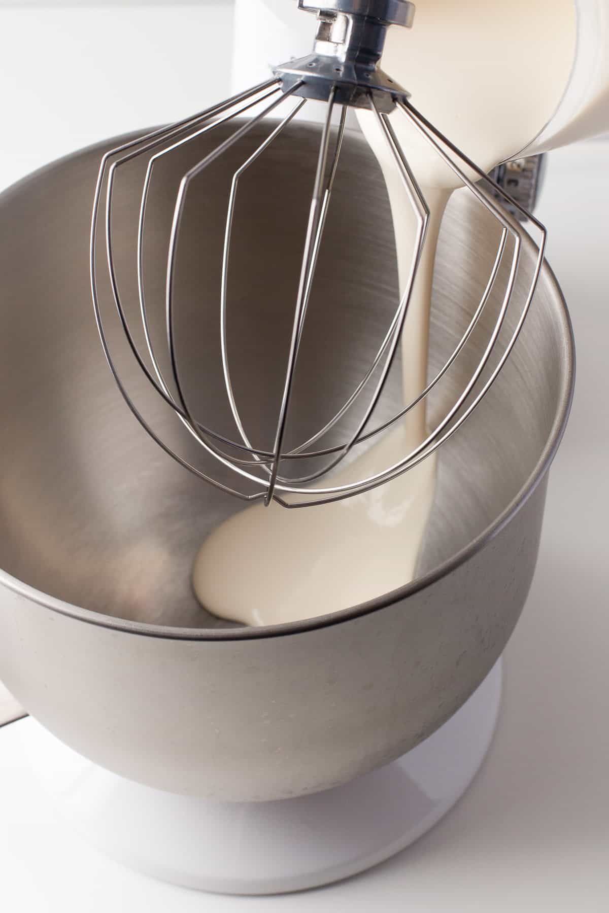 Heavy cream being poured into a metal bowl of a stand mixer with a whisk attachment.