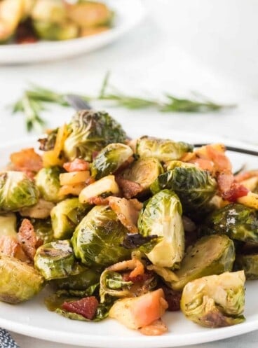 A mound of roasted brussels sprouts with bacon and apples o n a white plate with rosemary in the background.