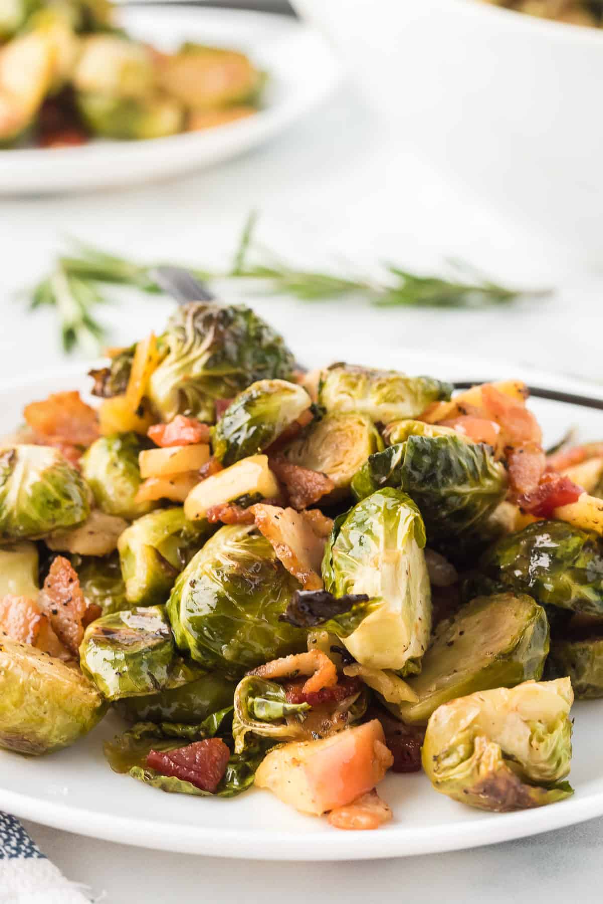 A mound of roasted brussels sprouts with bacon and apples o n a white plate with rosemary in the background.
