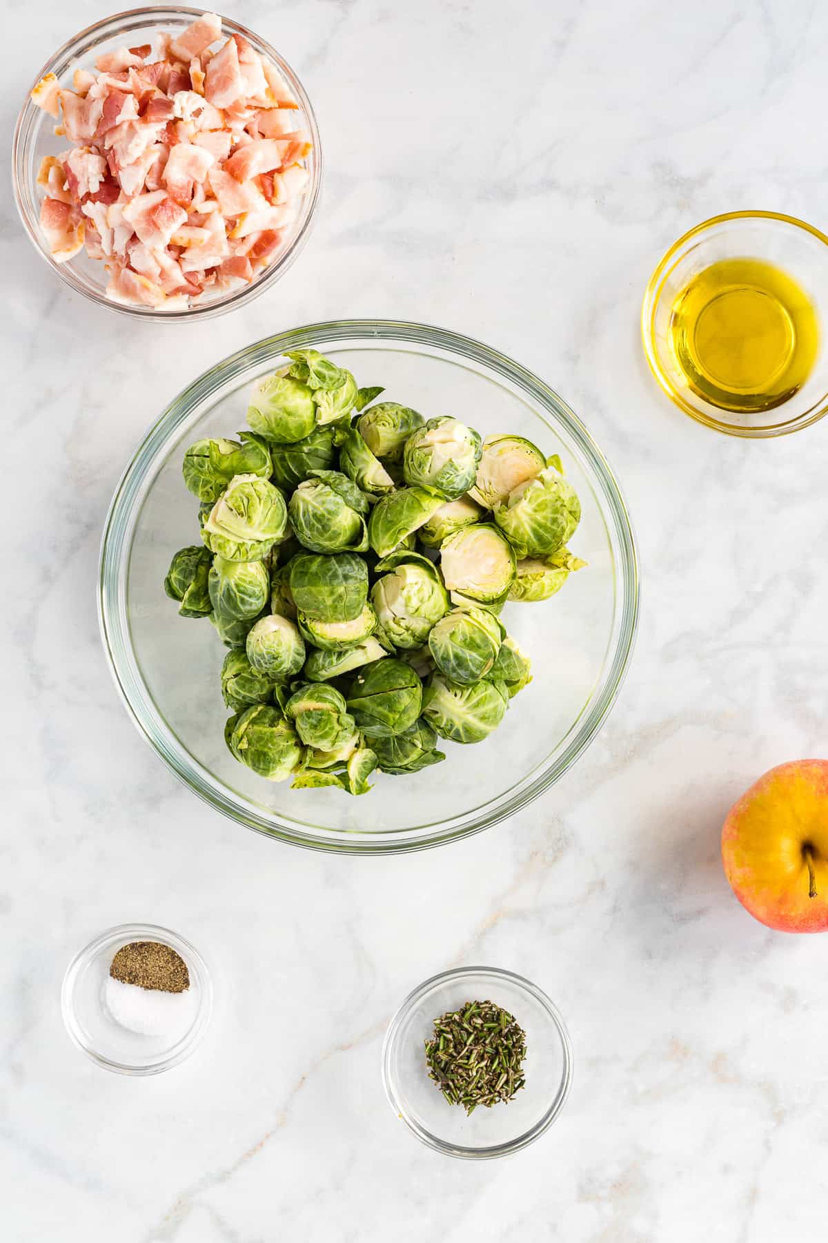 Bowls of brussels sprouts, chopped bacon, olive oil, seasonings, and an apple on a white surface.