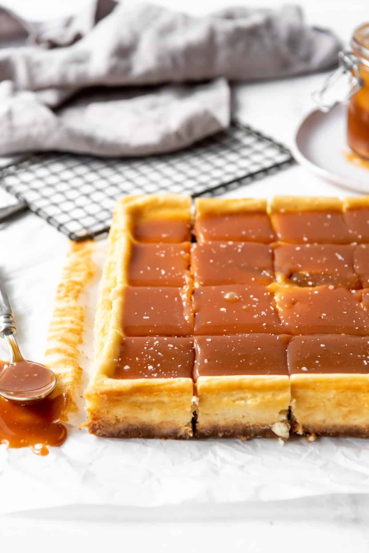 Cut cheesecake bars covered in caramel sauce and sprinkled with salt flakes are resting on white parchment paper, with a jar of caramel and a wire cooling rack in the background. 