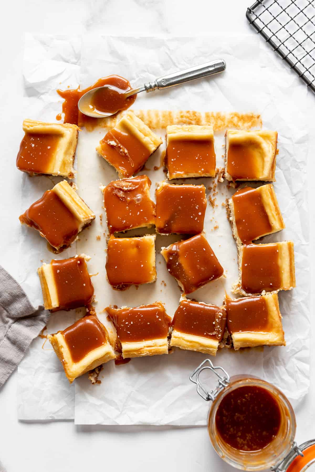 squares of cheesecake covered in salted caramel on white parchment paper next to a jar of homemade caramel sauce.