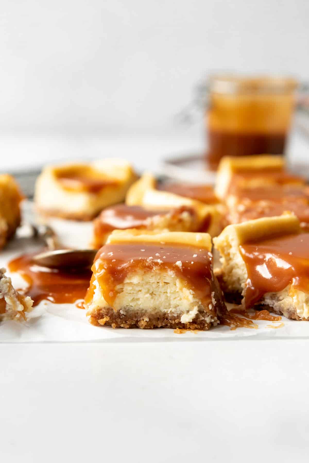 Side profile of squares of cheesecake bars covered in caramel sauce scattered on white parchment paper. 