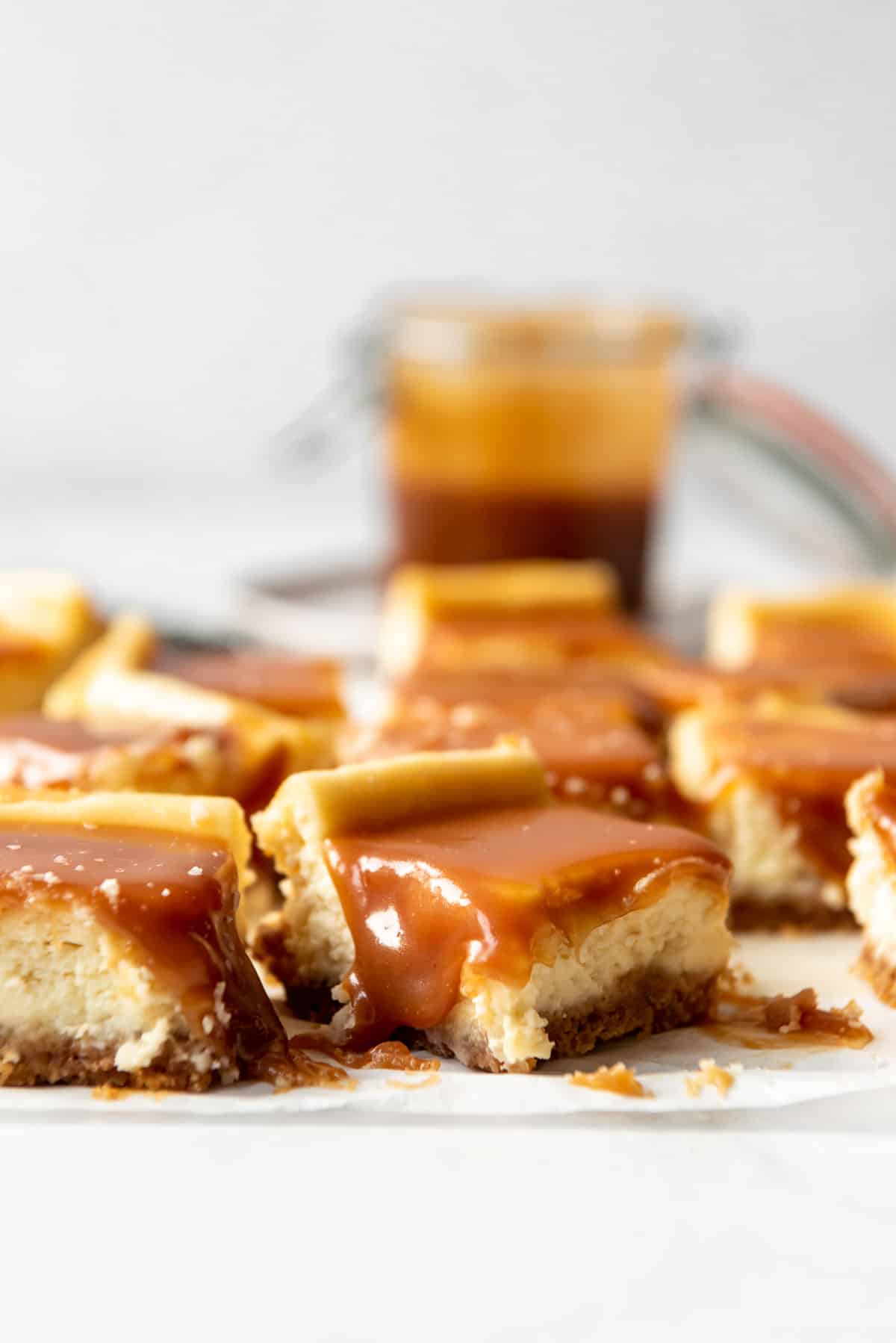 Cut cheesecake bars topped with caramel sauce are arranged on parchment, in a close up side view showing a crust, filling and topping. 