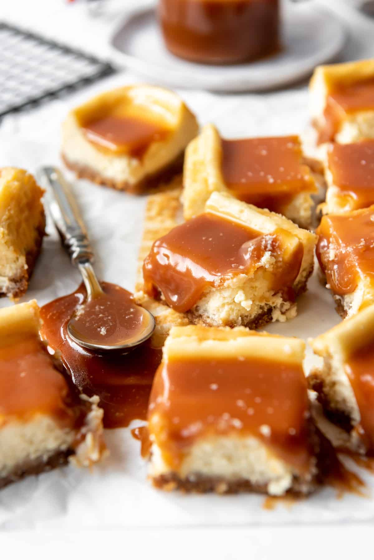 Squares of salted caramel cheesecake bars are scattered on white parchment, near a spoon covered in caramel sauce. A jar of sauce and a wire cooling rack are visible in the background. 