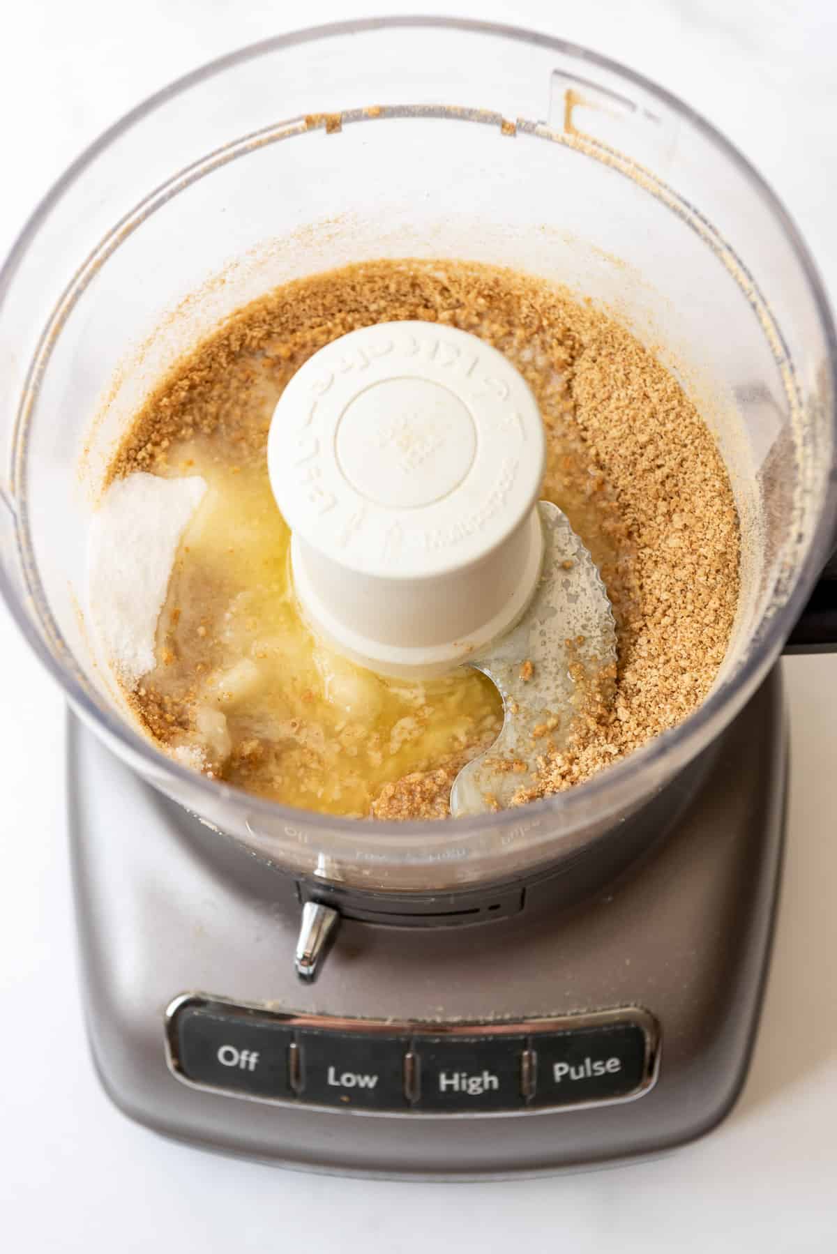 Crushed graham crackers, melted butter and granulated sugar are in a food processor, not yet blended together. 