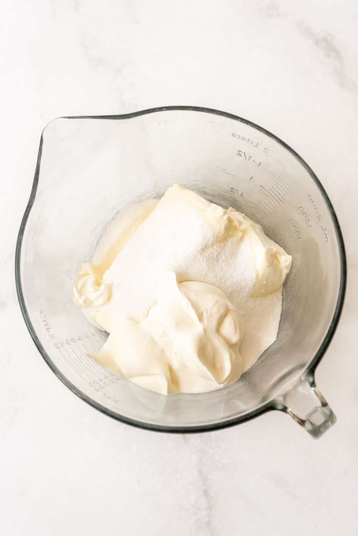 Softened cream cheese is in a glass mixing bowl, with granulated sugar and sour cream added in clumps on top. 