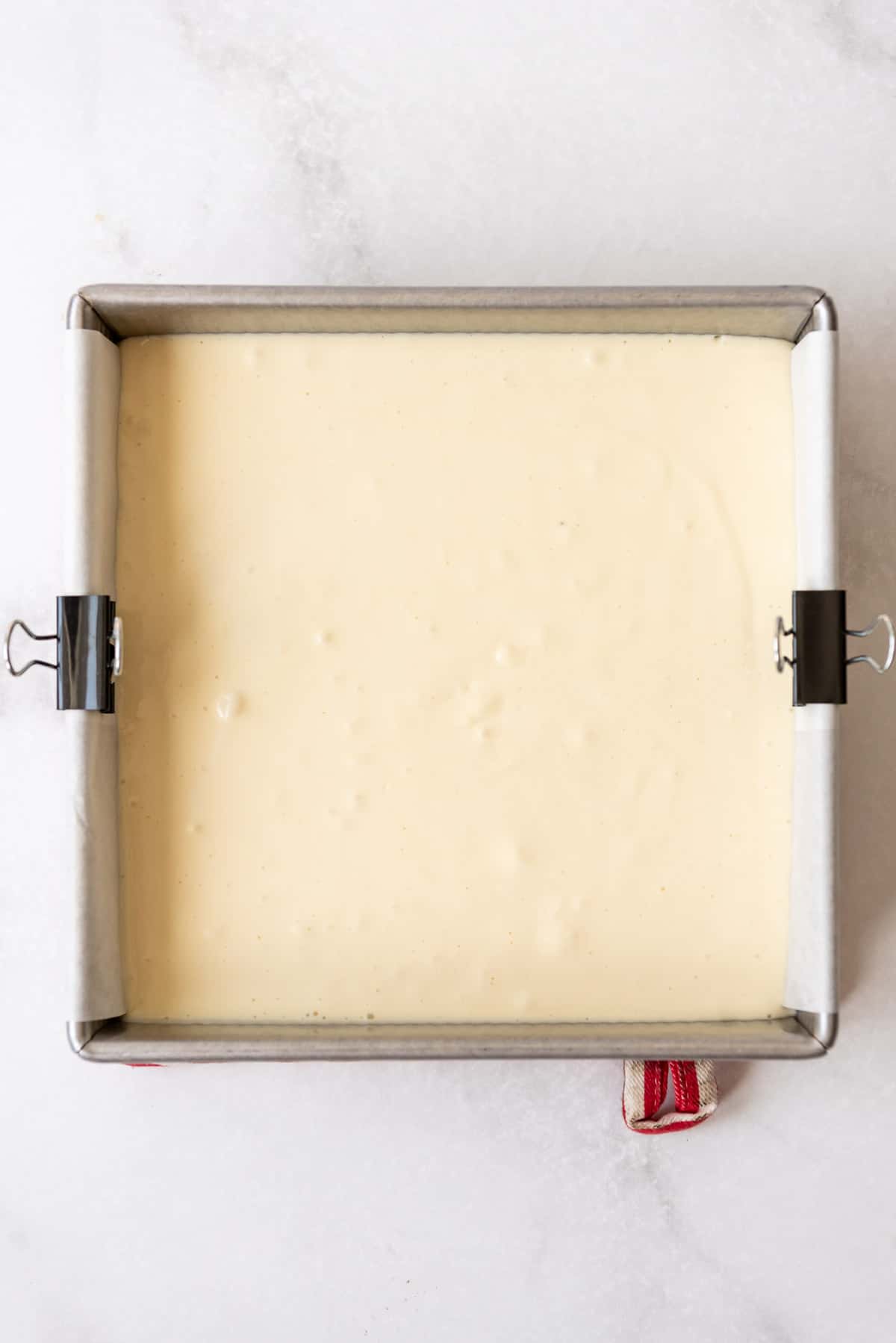 Blended cheesecake filling has been poured on top of the partially baked crust in a metal pan with a parchment paper sling visible on the sides. 