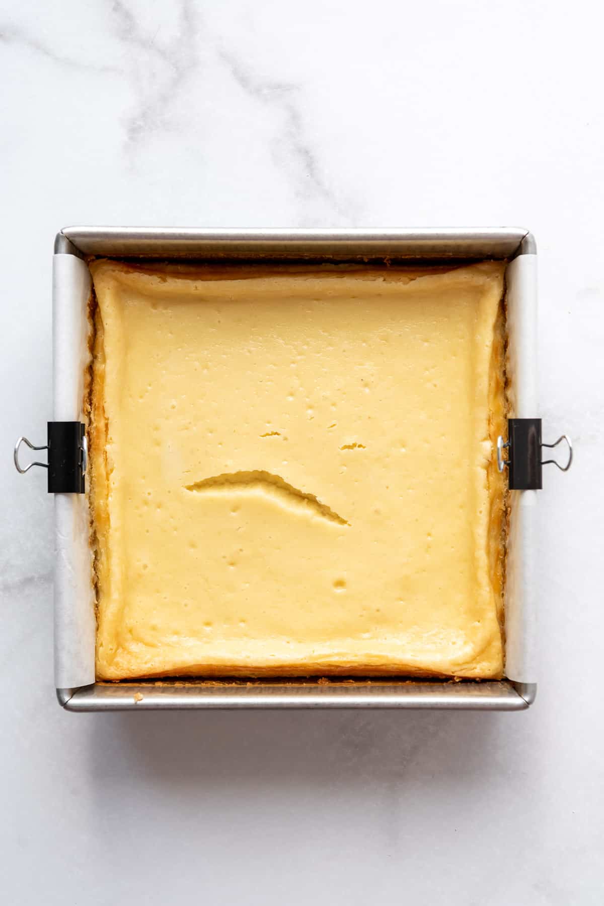 Baked cheesecake is resting on a countertop in a square metal pan. 