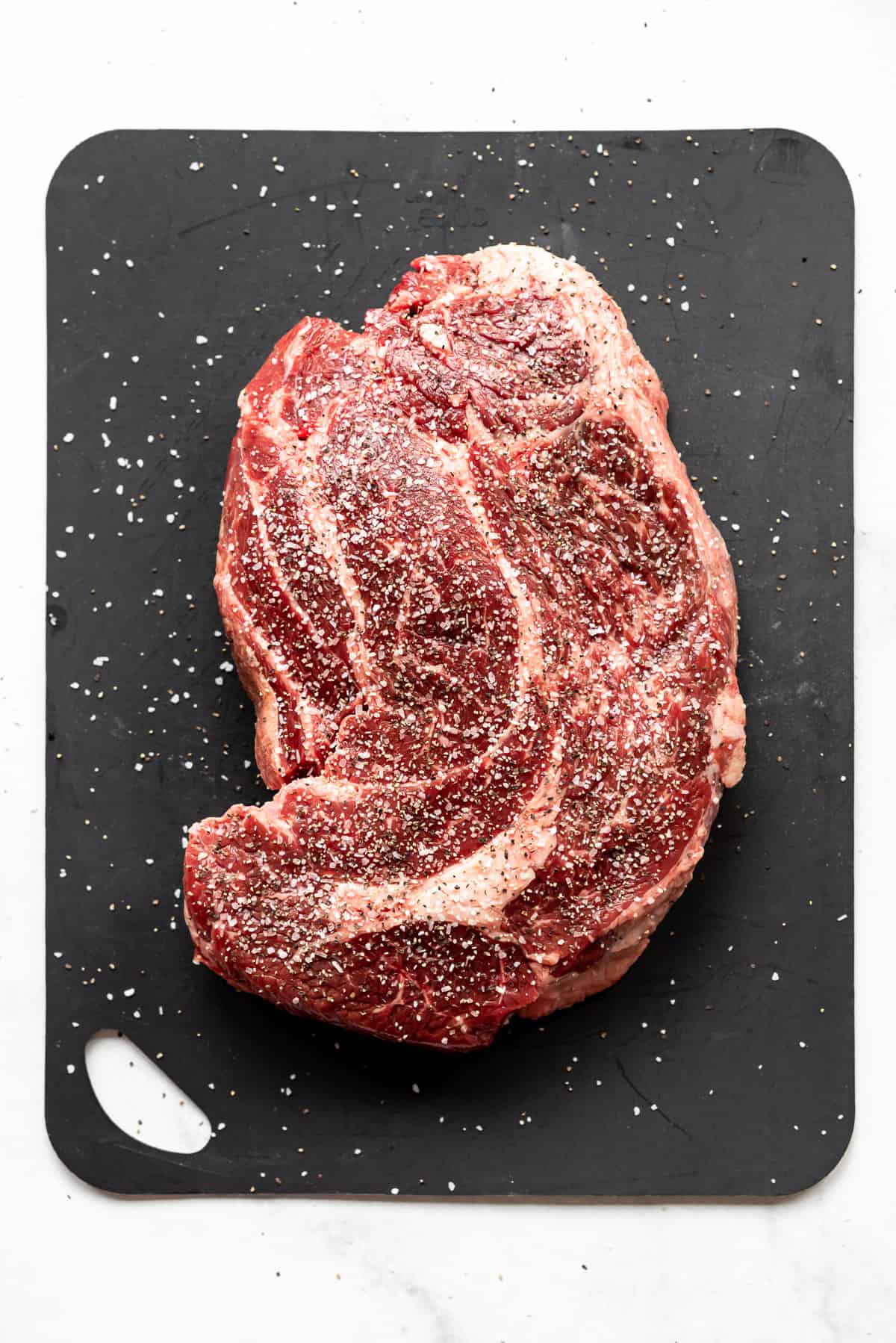 A chuck roast has been seasoned with salt and pepper and is resting on a black cutting board. 