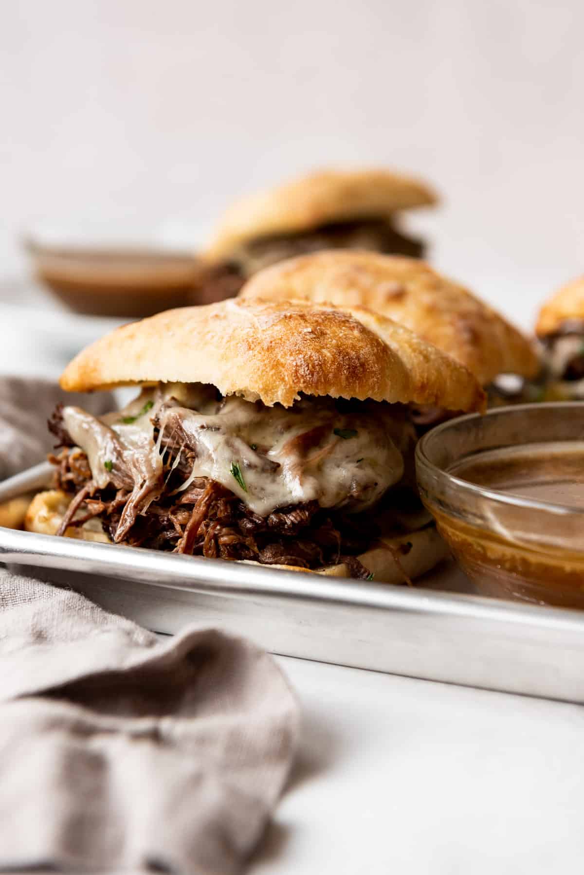 A shredded beef sandwich with melted cheese is visible next to a bowl of au jus, resting on a silver tray. Other sandwiches and another bowl are visible in the background. 