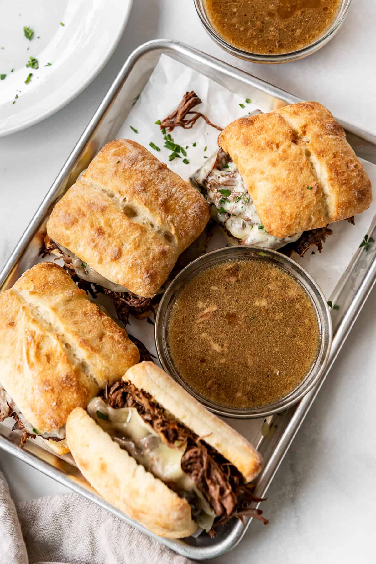 French dip sandwiches are displayed on a metal tray with a bowl of au jus to dip. 