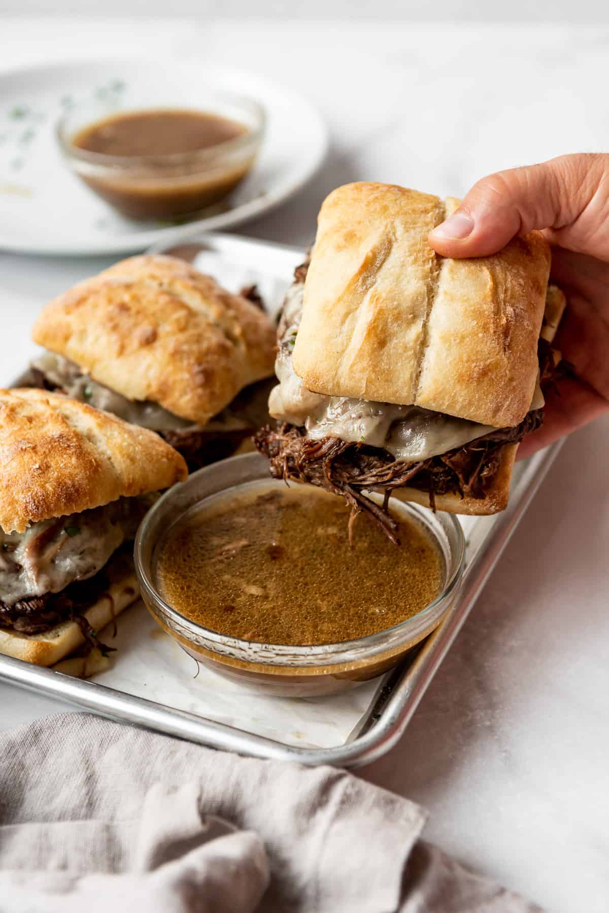 A hand holding a french dip sandwich on a ciabatta roll above a clear bowl containing au jus. 