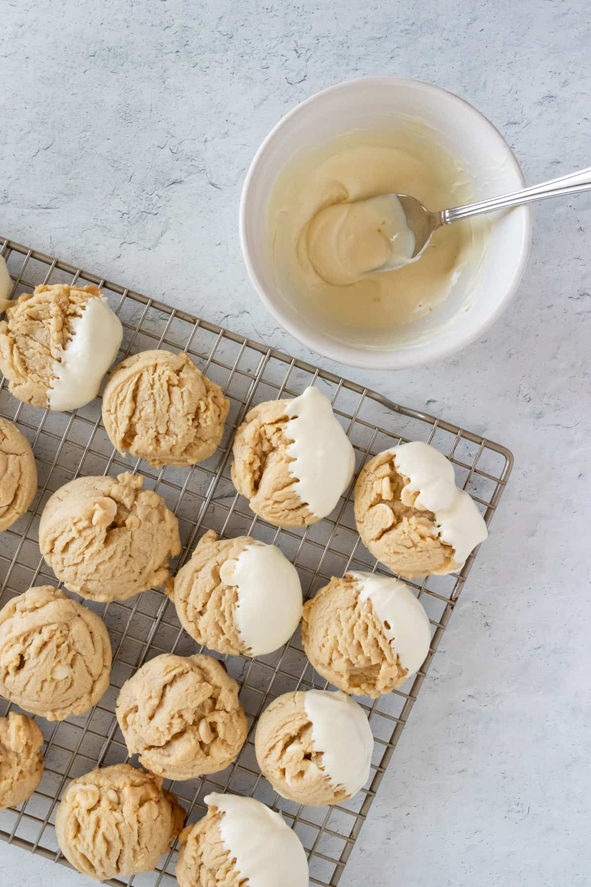 Peanut butter white chocolate chip cookies are cooling on a wire rack. Seven of the cookies are partially dipped in white chocolate, and a small bowl of melted white chocolate is displayed next to the wire rack. 