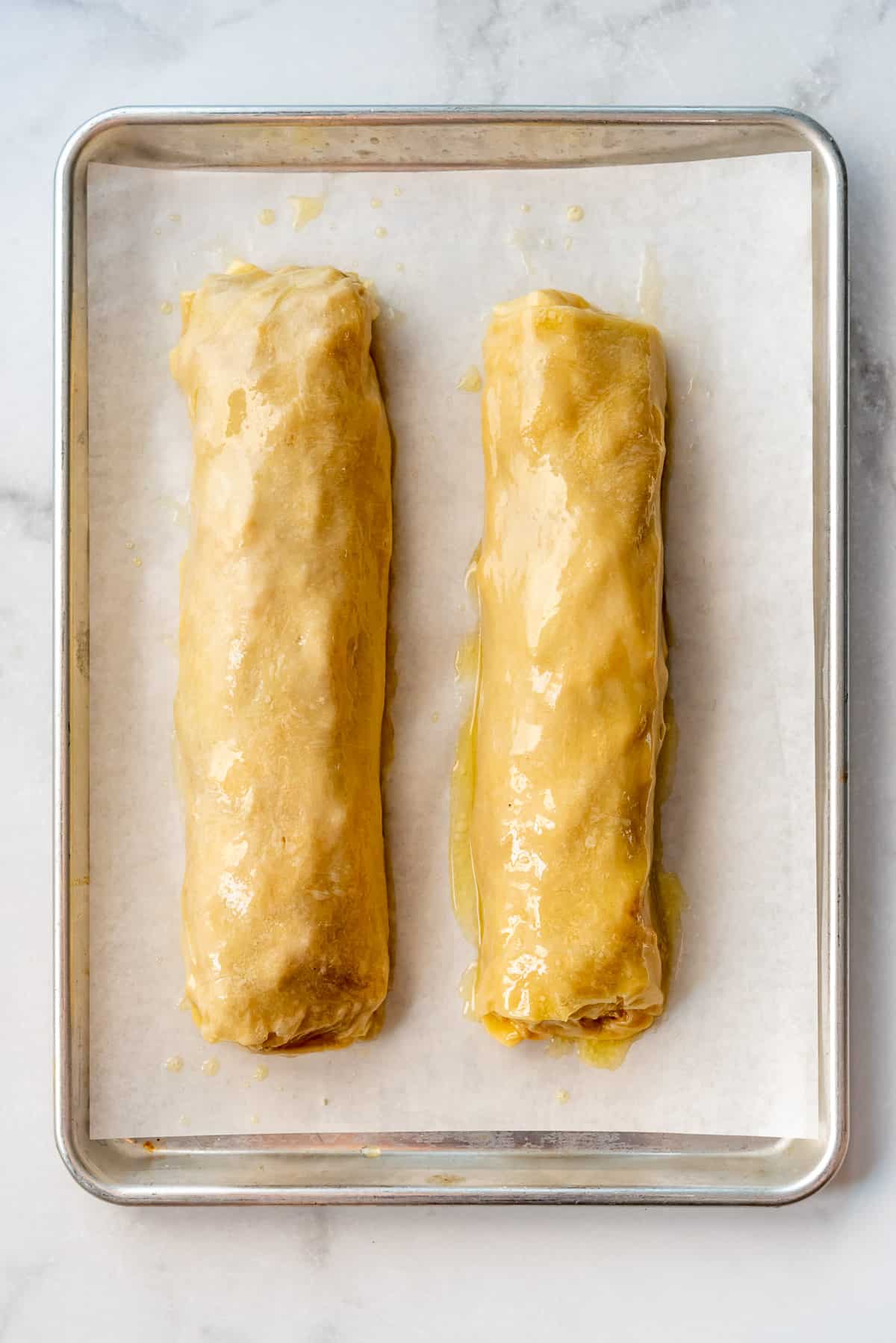 two apple strudel rolls brushed with melted butter on a baking sheet lined with parchment paper.