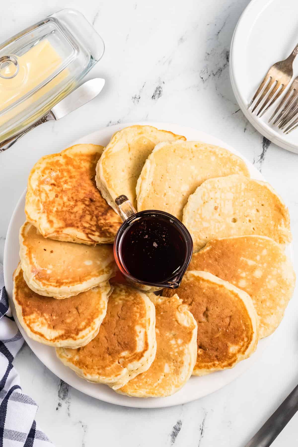 A plate of fluffy buttermilk pancakes around a small glass pitcher of maple syrup.