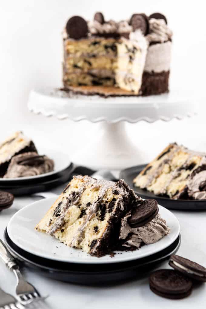 A slice of Oreo cake with cookies and cream frosting on a white plate in front of the rest of the cake.