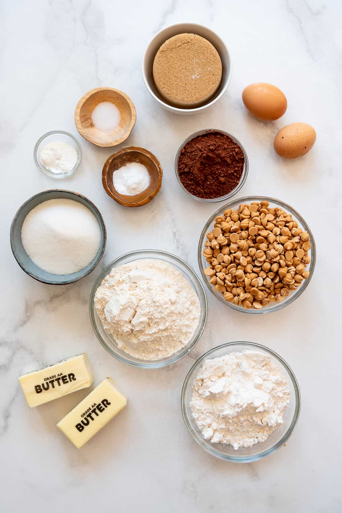 Ingredients for making chocolate cookies with peanut butter chips in separate bowls on a white surface.