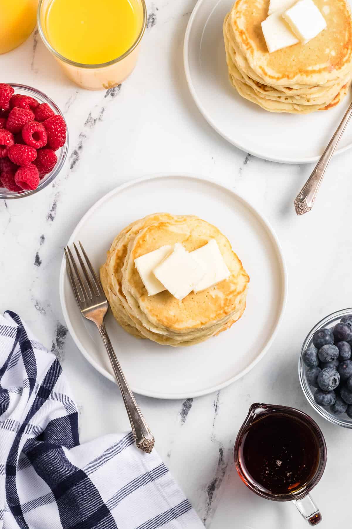 Two white plates with stacks of pancakes topped with pats of cold butter with orange juice, berries, and syrup beside them.