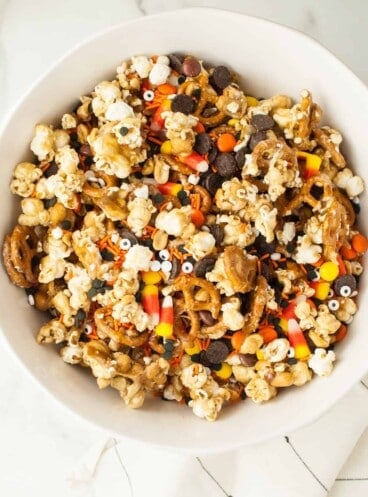 A large white bowl filled with caramel corn, candy corn, pretzels, chocolate, and peanuts.
