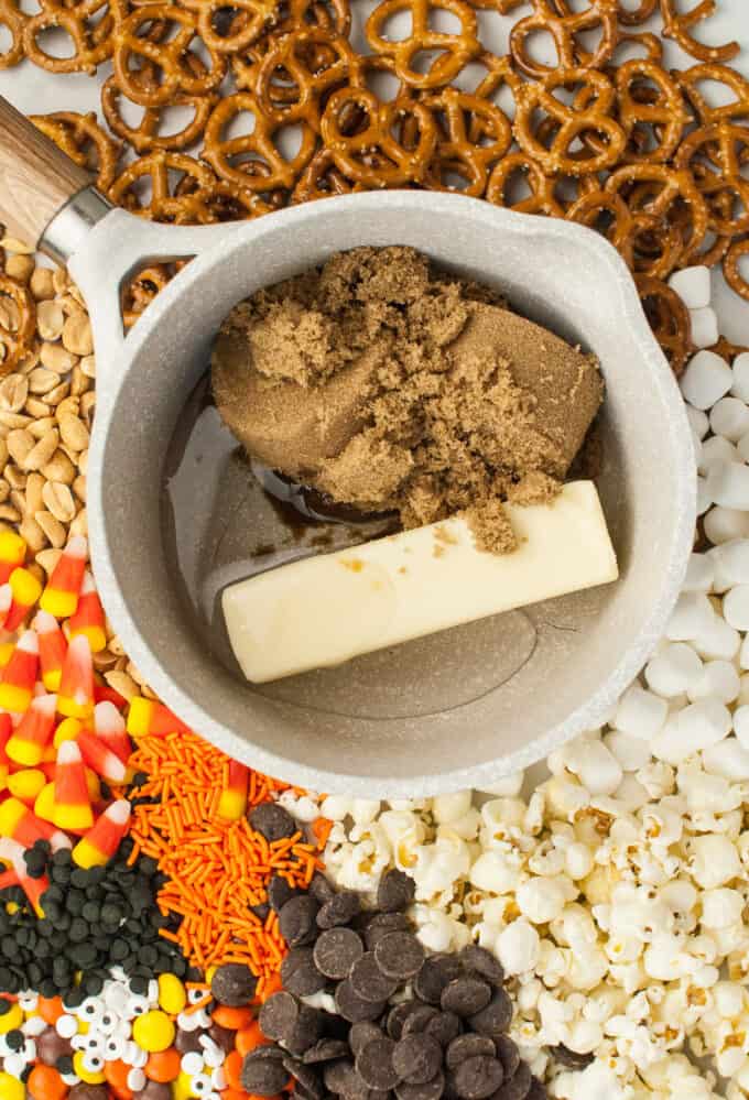 Butter, brown sugar, and corn syrup in a pot surrounded by popcorn, sprinkles, peanuts, chocolate, and candy corn.