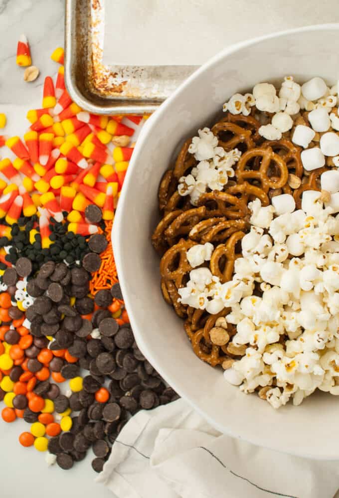 Popcorn, pretzels, and peanuts in a bowl with candy corn and chocolate chips next to it.