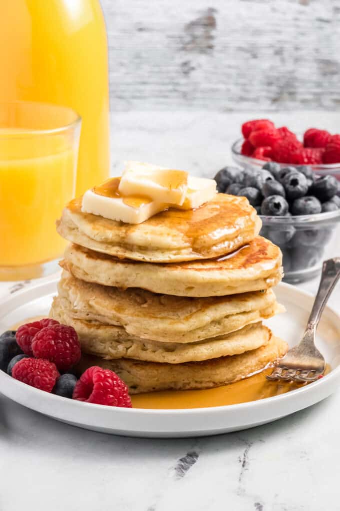Pancakes on a plate with syrup and fresh fruit in front of a pitcher of orange juice.