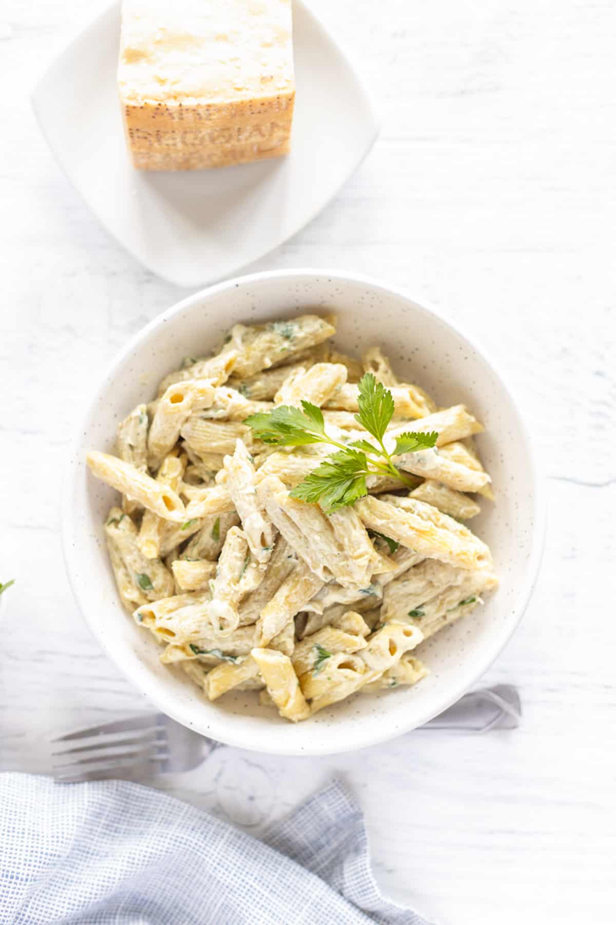 A white bowl of Instant Pot Creamy Penne Alfredo topped with fresh parsley and a parmesan cheese wedge on the side