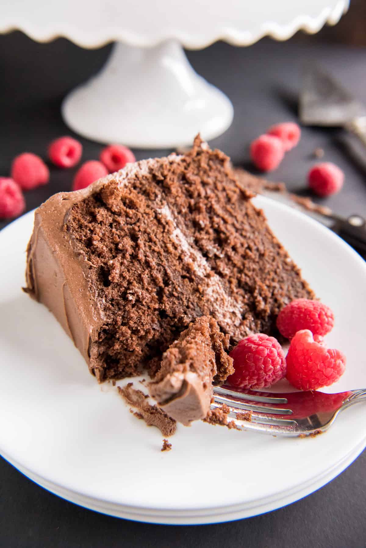 A slice of chocolate cake on a white plate missing a bite and next to fresh raspberries.