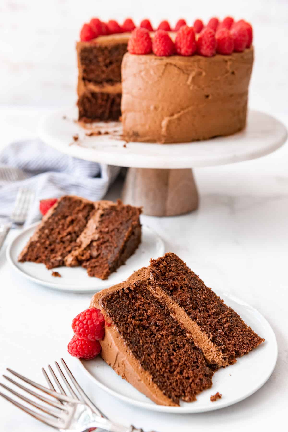 A slice of chocolate cake removed from the whole and placed on a white plate below a cake stand.