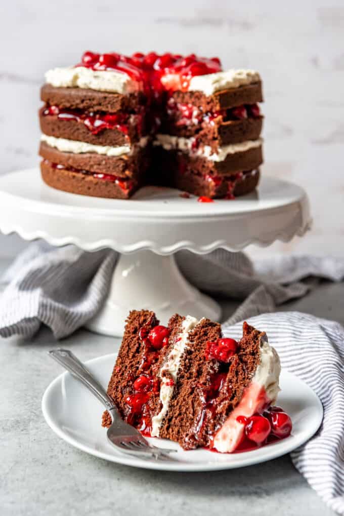 A black forest cake on a cake stand with a slice removed and placed on a plate below.