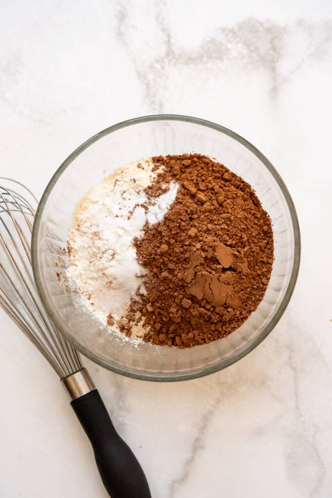 Flour, cocoa powder, baking powder, baking soda, and salt in a bowl next to a whisk.
