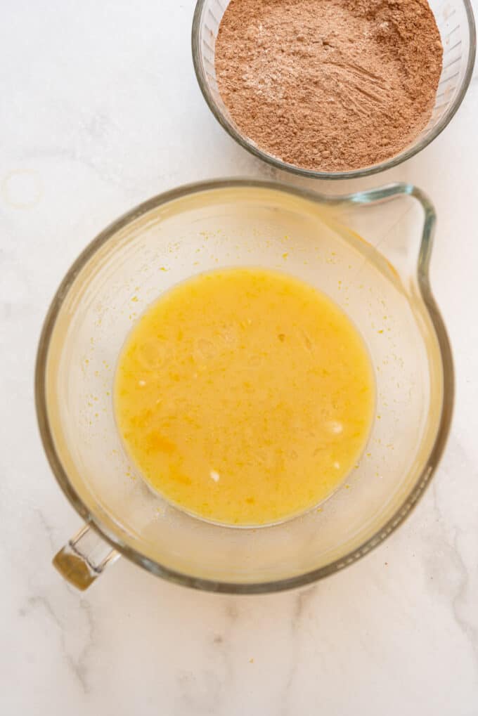 Mixed sugar, eggs, oil, and vanilla extract in a glass bowl with a bowl of whisked dry ingredients nearby.