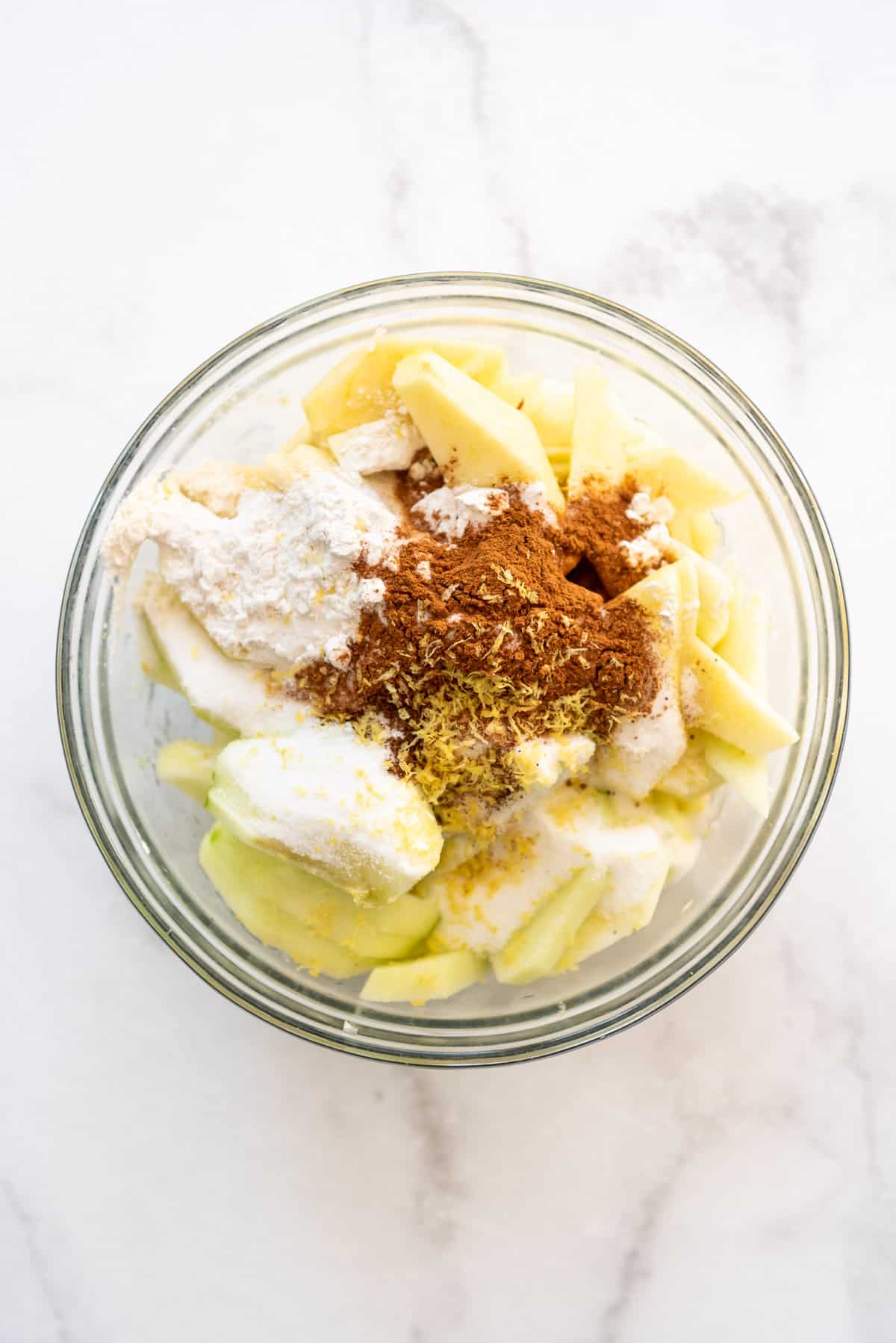 Sliced apples in a large glass bowl with flour, sugar, cinnamon, and lemon zest.