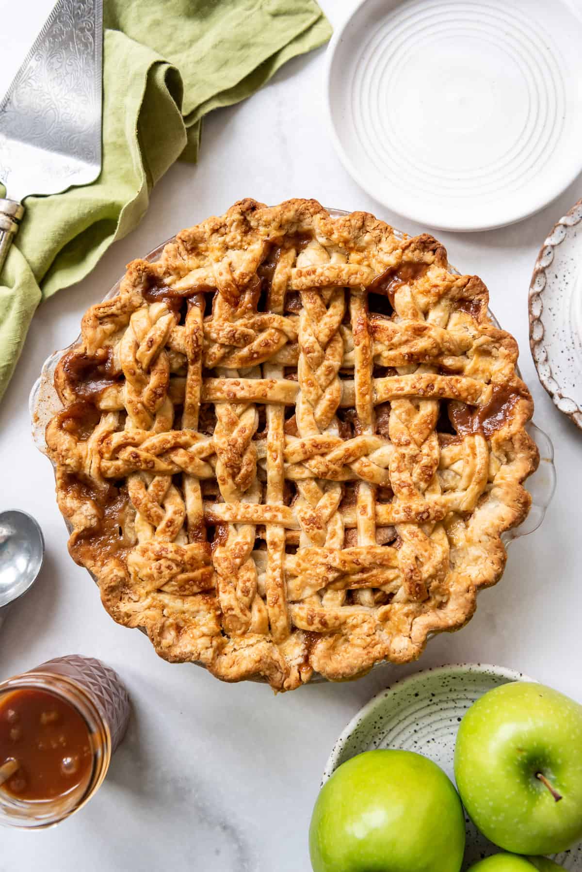 An apple pie with a braided lattice crust next to green granny smith apples and caramel sauce.