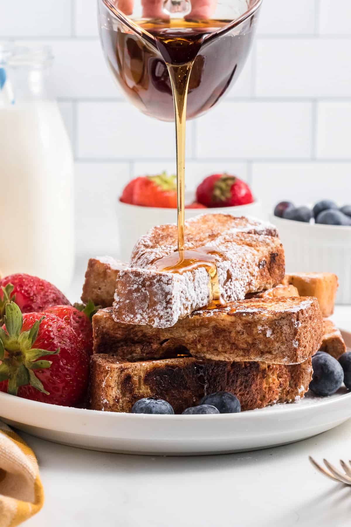 A pile of french toast sticks on a white plate topped with powdered sugar and garnished with fresh berries.