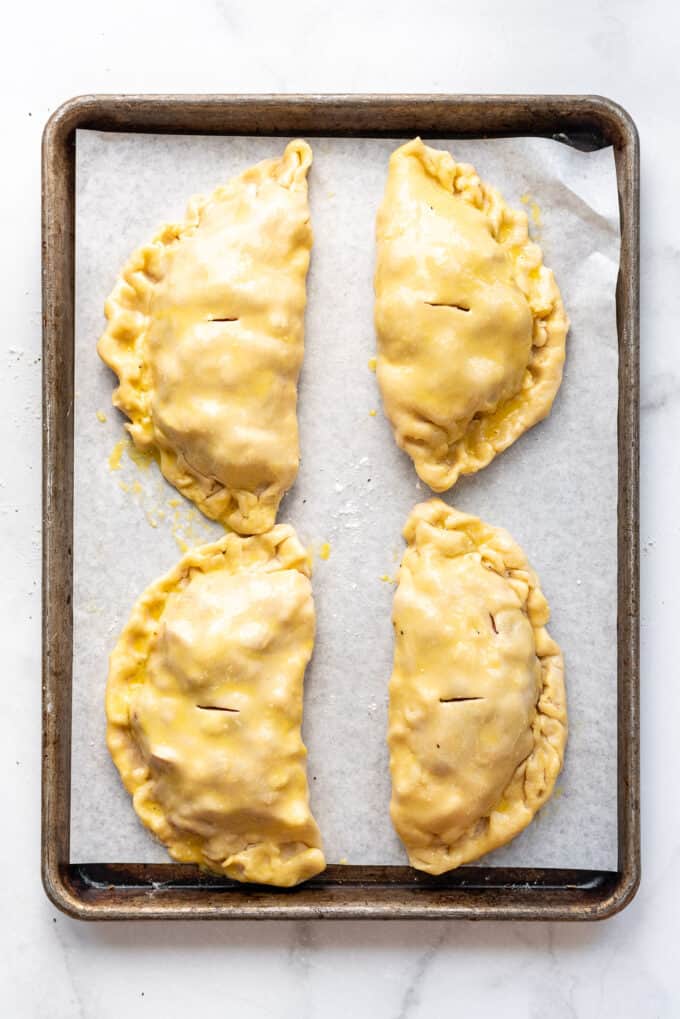 Four unbaked Cornish pasties brushed with egg wash on a baking sheet.