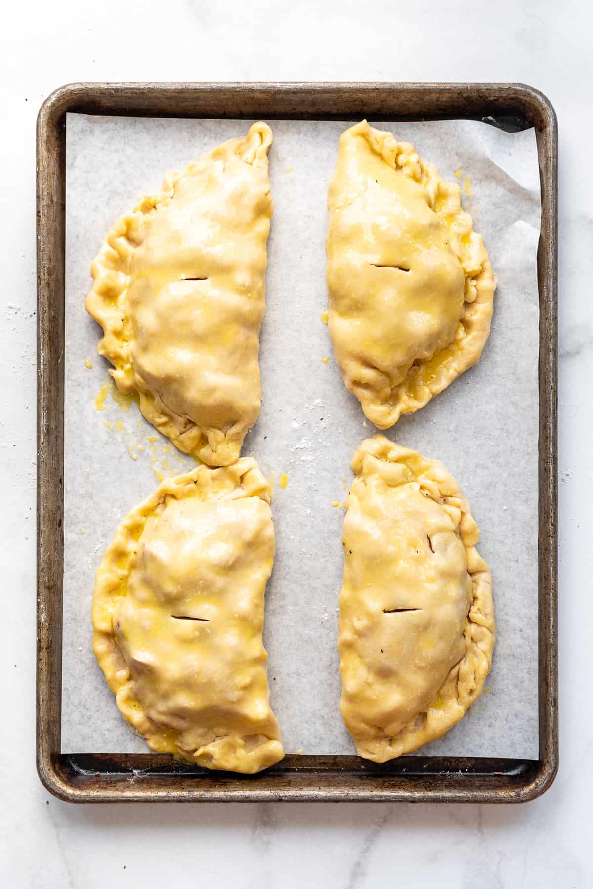 Four unbaked Cornish pasties brushed with egg wash on a baking sheet.